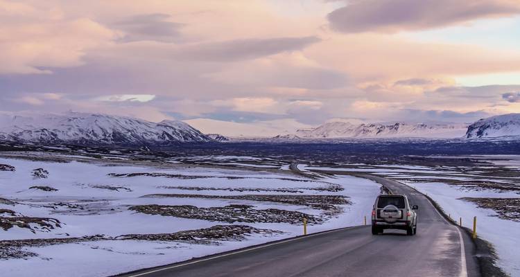 Snowy landscape with a car on a winding road and a colorful sky.