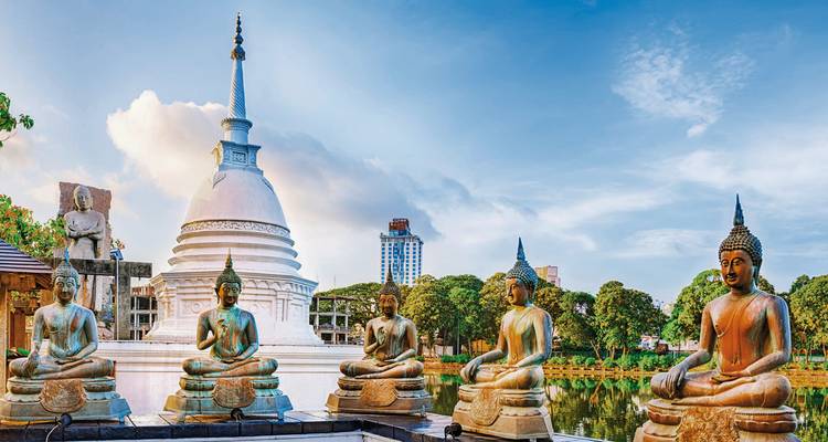 Row of bronze Buddha statues beside white stupa reflected in serene lake with city skyline beyond