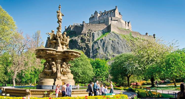 Castle on a rocky outcrop with a fountain and gardens in the foreground.