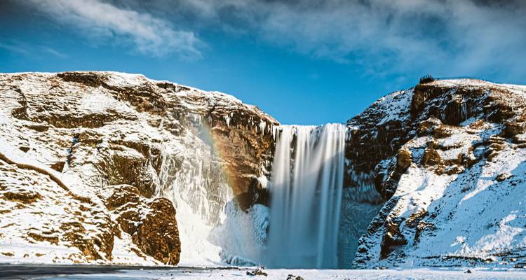 Krachtige Skógafoss waterval die stroomt tussen besneeuwde kliffen met een zichtbare regenboog.