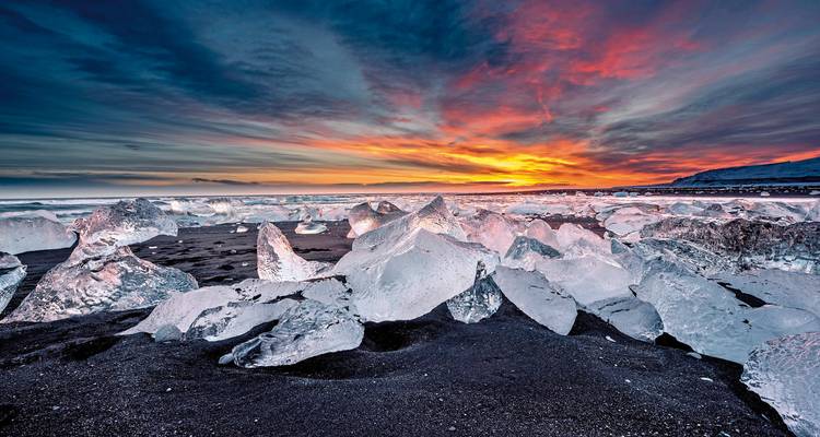 Glinsterende ijsfragmenten op zwart zand op Diamond Beach onder een vurige zonsonderganghemel.