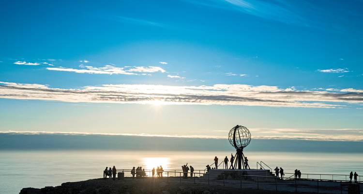 Atardecer junto a un acantilado con un monumento de globo terráqueo y turistas.