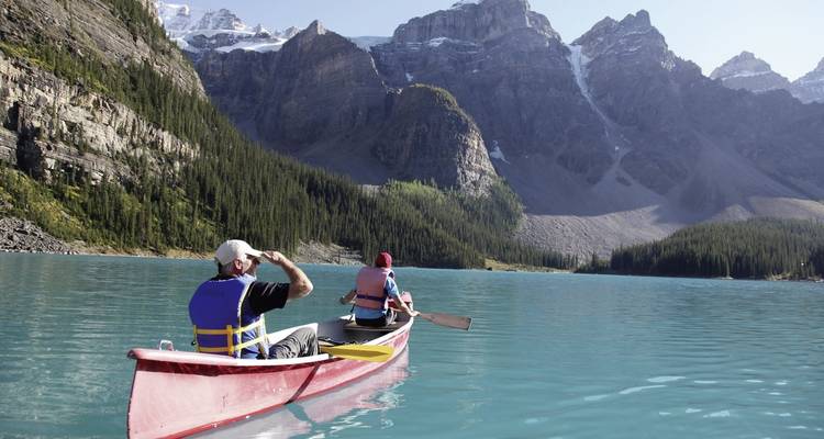 Zwei Menschen fahren Kanu durch einen türkisfarbenen See mit Bergen im Hintergrund.