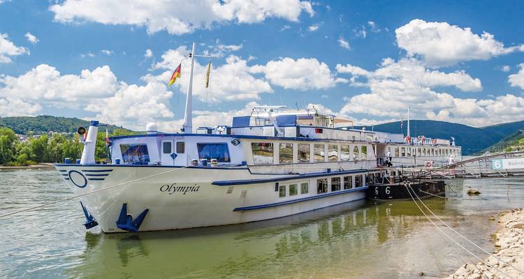 Navire de croisière amarré sur une rivière avec des drapeaux flottants et des collines en arrière-plan.