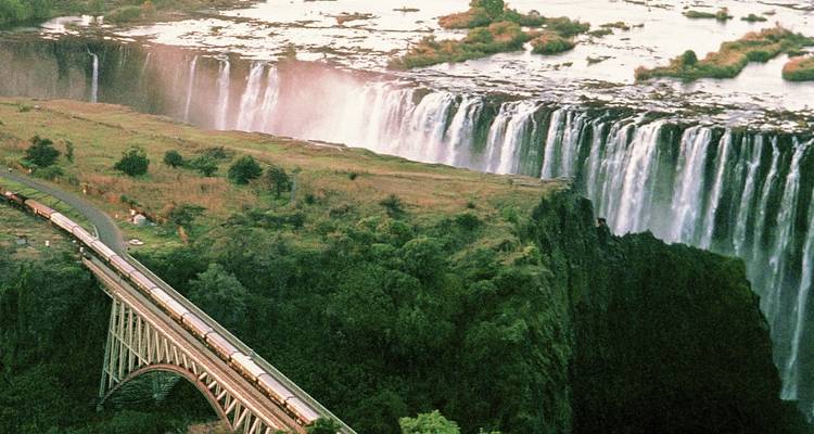 Train crossing a bridge over a waterfall in a lush landscape.