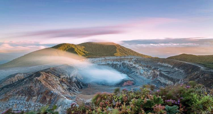 Crater with smoke and surrounding lush landscape under pink skies.