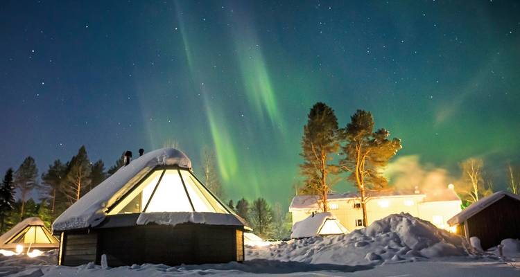 Nordlichter über schneebedeckten Hütten in einem bewaldeten Gebiet bei Nacht.