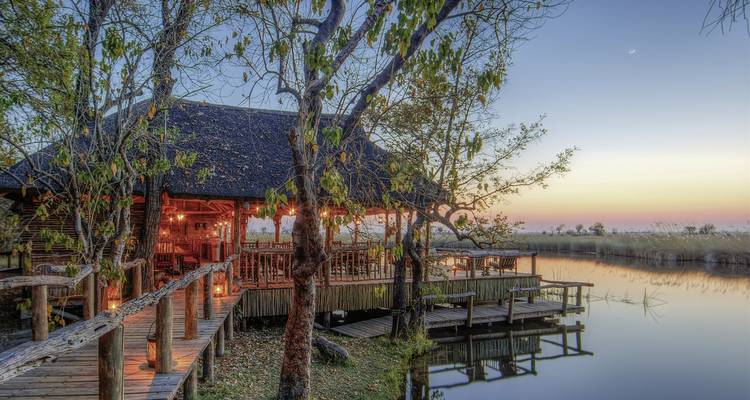 A serene riverside lodge with a wooden deck under a twilight sky, surrounded by trees and a river.