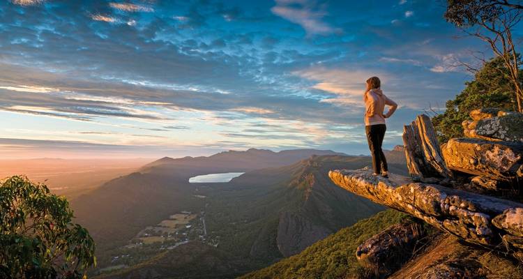 Une personne debout au bord d'une falaise surplombant une vaste vallée montagneuse au coucher ou au lever du soleil.