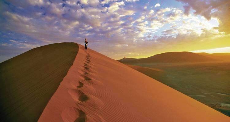 Person walking along the crest of a golden sand dune during sunset.
