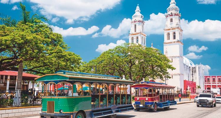 Colorful trams on a street with two towers and lush trees.