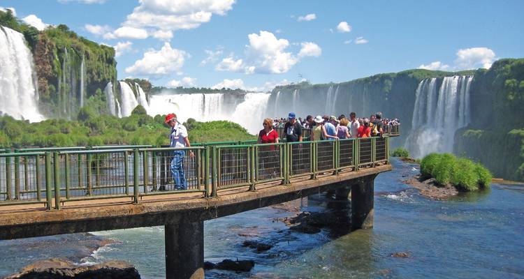 Touristen gehen auf einer Brücke mit Wasserfällen im Hintergrund.