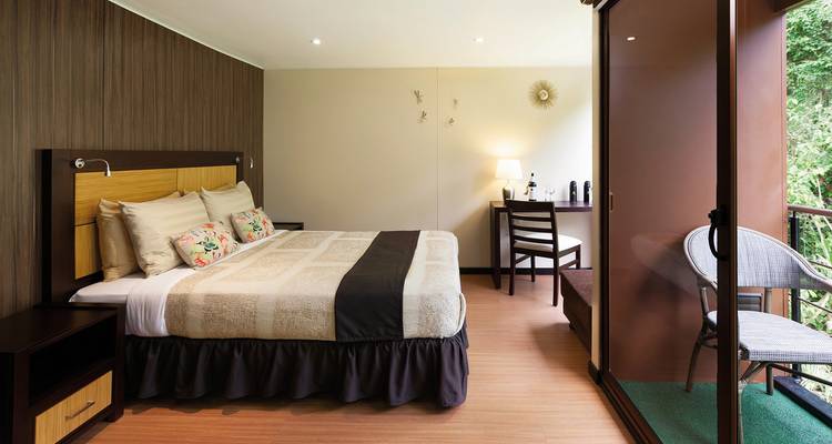 Chambre d'hôtel moderne avec un grand lit, un bureau et un balcon donnant sur une végétation luxuriante.