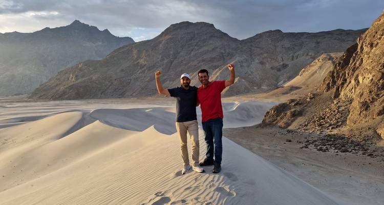Two friends celebrating on golden sand dunes with rugged mountains behind them.