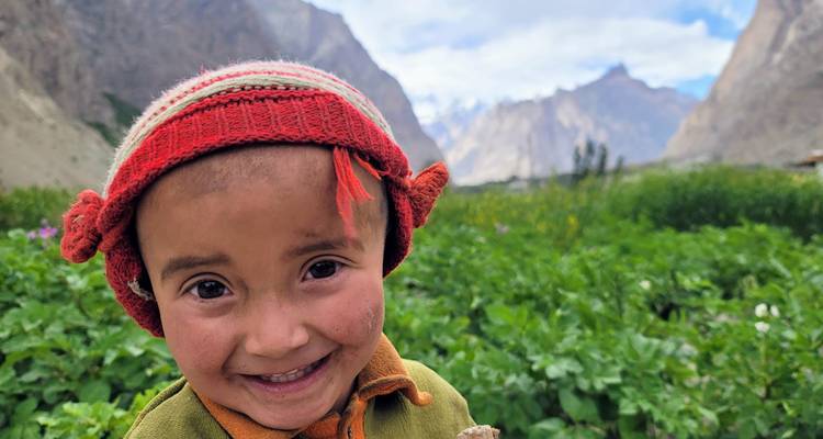 A cheerful child wearing a red wool hat standing in a lush green valley with jagged peaks beyond.