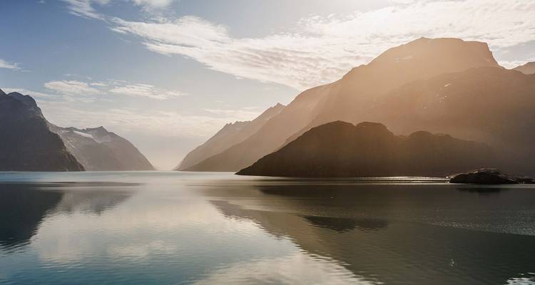 Soft sunlight filters through misty peaks over a glassy fjord, creating a perfect mountain reflection.