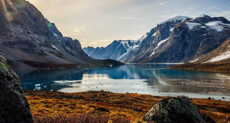 Dramatic snow-capped peaks surround a turquoise fjord dotted with small icebergs under golden light.