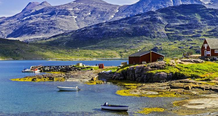 Small boats float by a rustic Greenlandic village set against green hills and towering peaks.