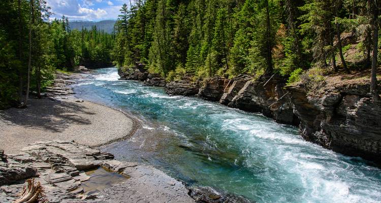 Ein klarer türkisfarbener Fluss stürzt durch eine felsige bewaldete Schlucht unter einem strahlend blauen Sommerhimmel.