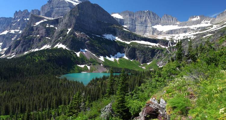 Vue panoramique de pics de granit parsemés de neige surplombant un lac de montagne turquoise et une vallée verdoyante luxuriante.