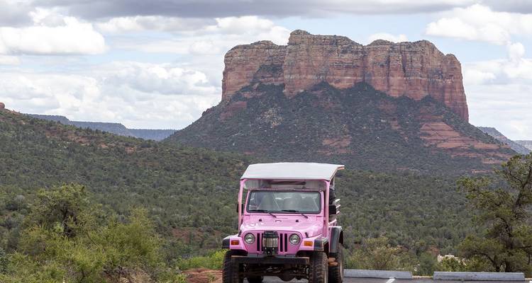Un jeep rose signature se tient sur un terrain de roche rouge avec les imposantes buttes de Sedona s'élevant derrière sous un ciel nuageux.