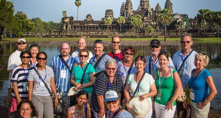 Large tour group smiles for a photo in front of Angkor Wat temple complex across a lotus pond.