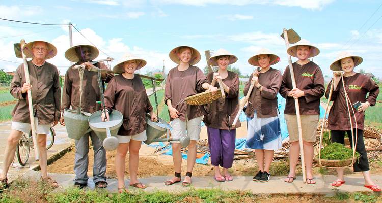 Smiling visitors in farming clothes and conical hats pose with tools in a vegetable field experience near Hoi An.