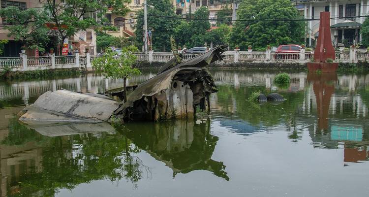 Wreckage of a downed military aircraft protrudes from a still urban lake surrounded by colonial-era buildings and greenery in the city.