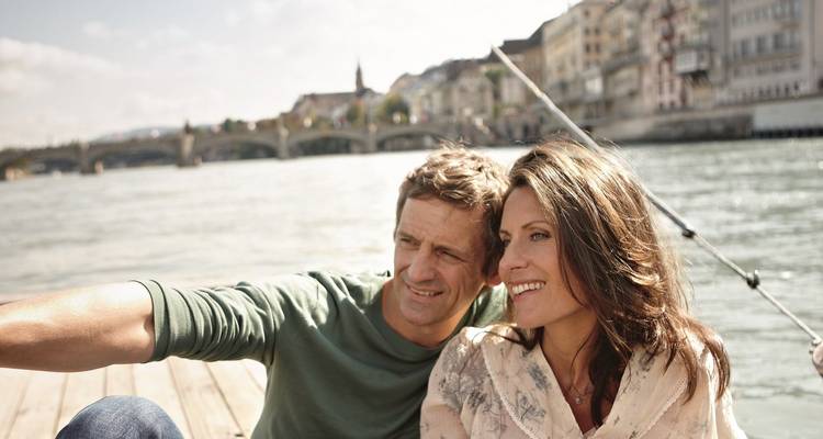 Una pareja se relaja en un muelle de madera junto a un amplio río europeo con edificios históricos de fondo.
