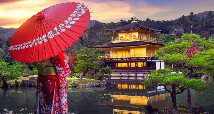 Une personne vêtue d'un kimono aux couleurs éclatantes tenant un parasol rouge contemple le temple doré Kinkaku-ji en bordure d'un étang reflétant les teintes du coucher de soleil