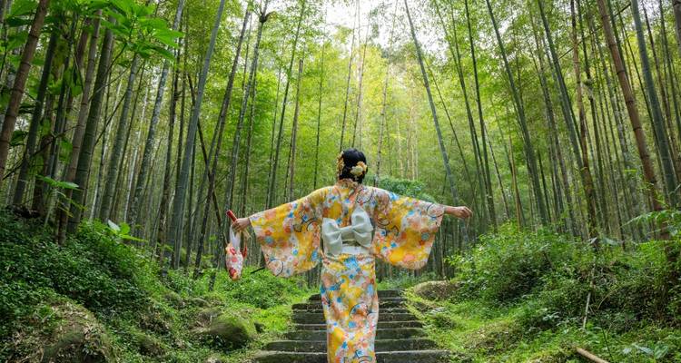 Femme en kimono floral montant les marches en pierre à travers une forêt de bambou imposante