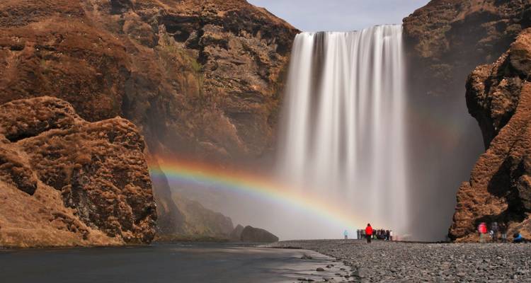 De machtige Skogafoss waterval stort zich over mosachtige rotsen met een levendige regenboog die zich boven de toeschouwers aan de voet uitstrekt.