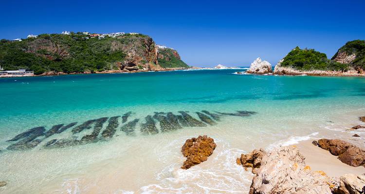 Baie turquoise cristalline aux Knysna Heads bordée de falaises rocheuses et de collines boisées.