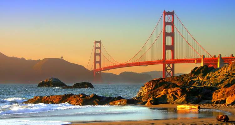 Le Golden Gate Bridge brille au coucher du soleil au-dessus des vagues de l'océan et des rochers accidentés du littoral.