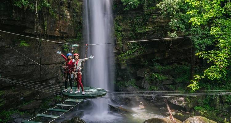 Abenteurer in Helmen stehen auf einer Holzplattform, die durch Kabel neben einem hohen Dschungelwasserfall aufgehängt ist.