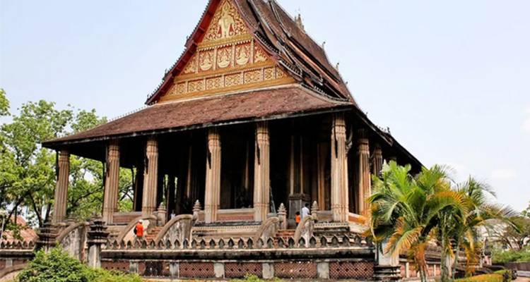 Der historische Wat-Si-Saket-Tempel mit hölzernen Säulen und gestuftem Dach liegt inmitten von Grünflächen unter einem blassen Himmel.