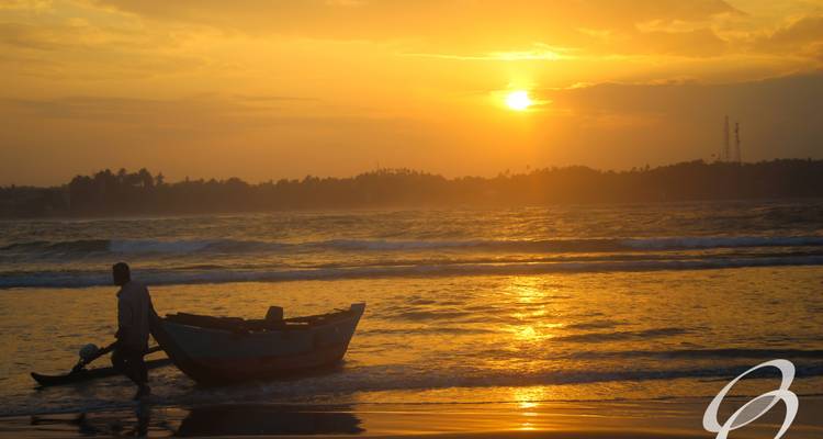 Een persoon die bij zonsondergang op het strand loopt met een kleine boot.