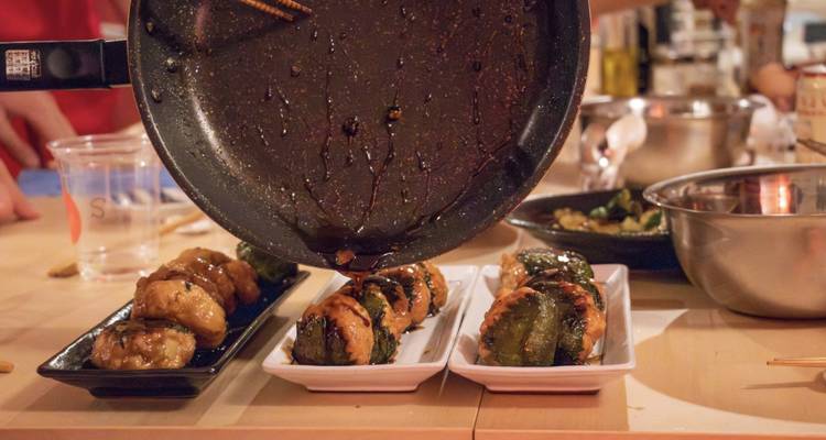 Cooking class scene where a pan drizzles sauce over neatly arranged skewered bites on plates.