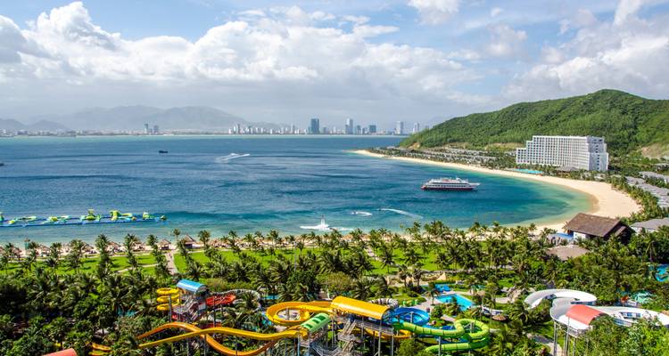 Luftaufnahme eines Wasserparks mit Rutschen und einem Strand in einer Bucht, mit einer Stadtsilhouette und Bergen im Hintergrund.