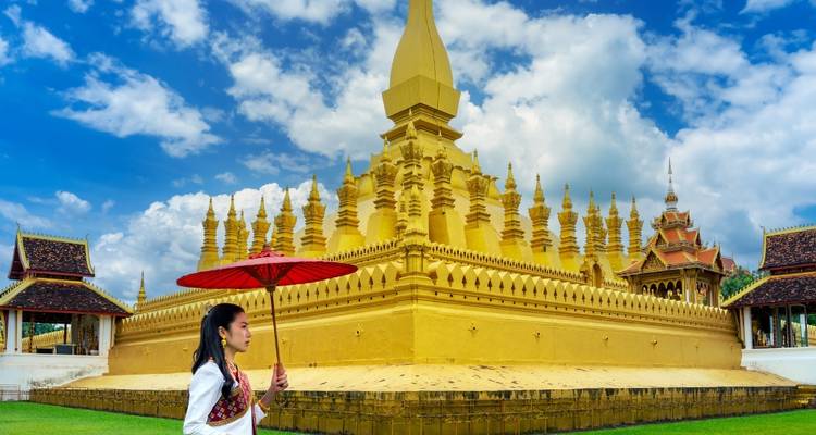 Femme vêtue d'une robe traditionnelle avec un parapluie rouge qui marche devant le stupa doré de Pha That Luang
