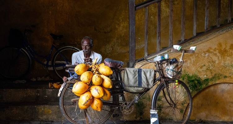 Ein Mann, der neben einem mit Kokosnüssen beladenen Fahrrad unter einer Treppe sitzt.