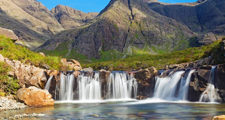 Clear mountain stream cascading over rocks in a series of small waterfalls with rugged peaks behind.