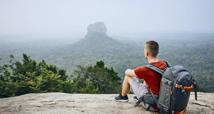 Backpacker zit op een rotsachtige richel met uitzicht op de mistige jungle en de iconische Sigiriya-rots die in de verte uittorent.