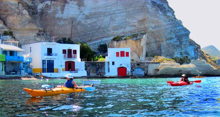 Twee mensen die kajakken in helder water met kleurrijke huizen tegen een rotsachtige achtergrond.