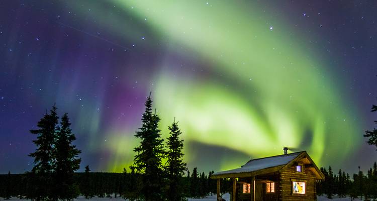 Vivid green and purple northern lights swirling above a cozy log cabin in snowy Alaska.