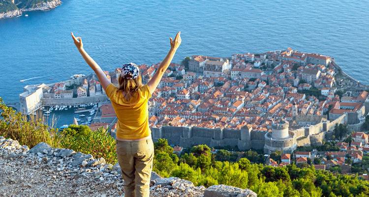 Voyageur levant joyeusement les bras en surplomb de la vieille ville fortifiée de Dubrovnik et de la mer Adriatique.