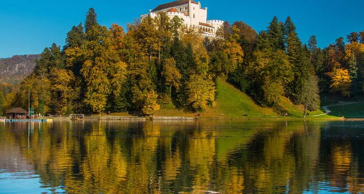 A white hill-top castle overlooks a calm lake surrounded by colorful autumn forest reflected in the water.