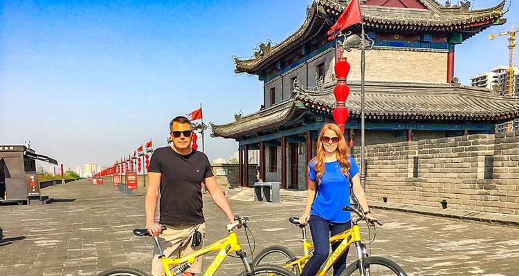 Couple posant avec des vélos sur un mur historique orné de drapeaux rouges.