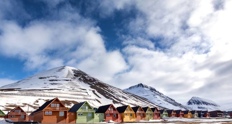 Una hilera de cabañas árticas de colores brillantes se alinea en la base de una montaña con vetas de nieve bajo un cielo dinámico.