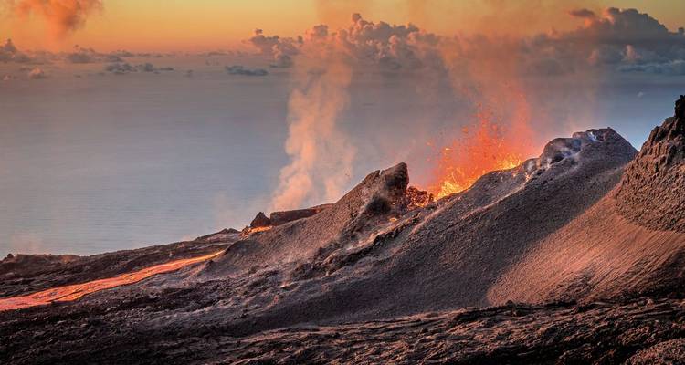 Éruption spectaculaire du volcan Piton de la Fournaise qui projette de la lave et des cendres sur fond de coucher de soleil dramatique sur l'océan